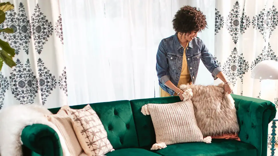 A woman standing behind a green couch with a bunch of frilly pillows.