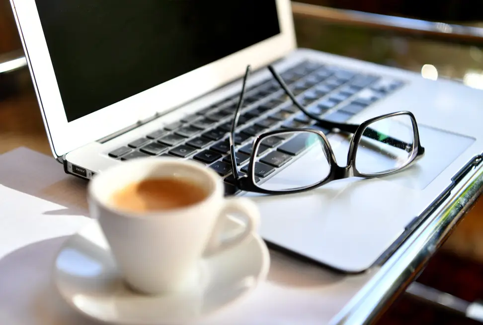 A laptop, hot beverage, and eyeglasses on a desk.