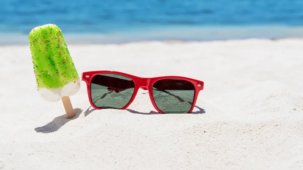 A pair of red Ray-Ban sunglasses lie on the sandy beach beside a green popsicle.