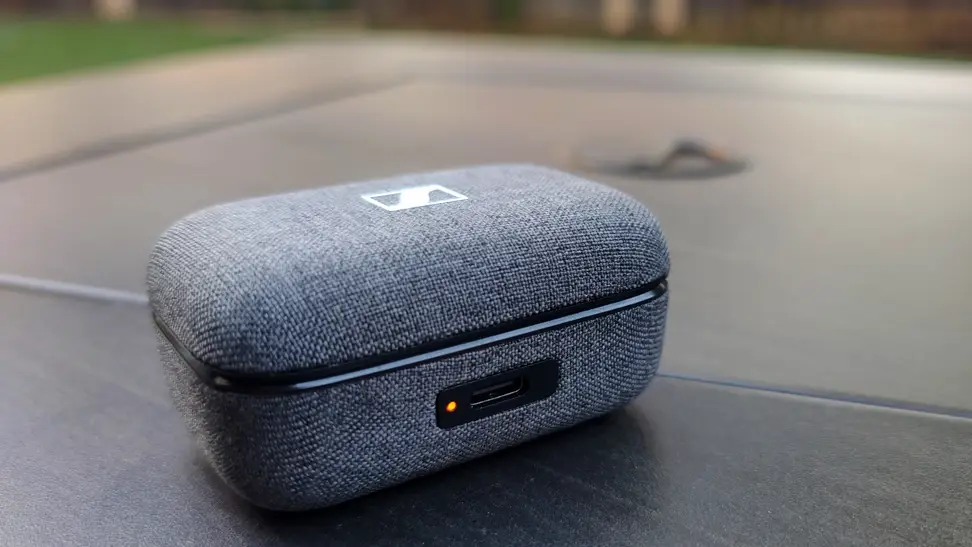 A gray fabric earbuds case sits on a black table with an orange LED at the front.
