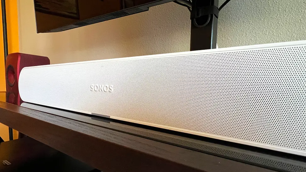 A white and tubular soundbar sits on a brown and black TV console with a TV mounted above.