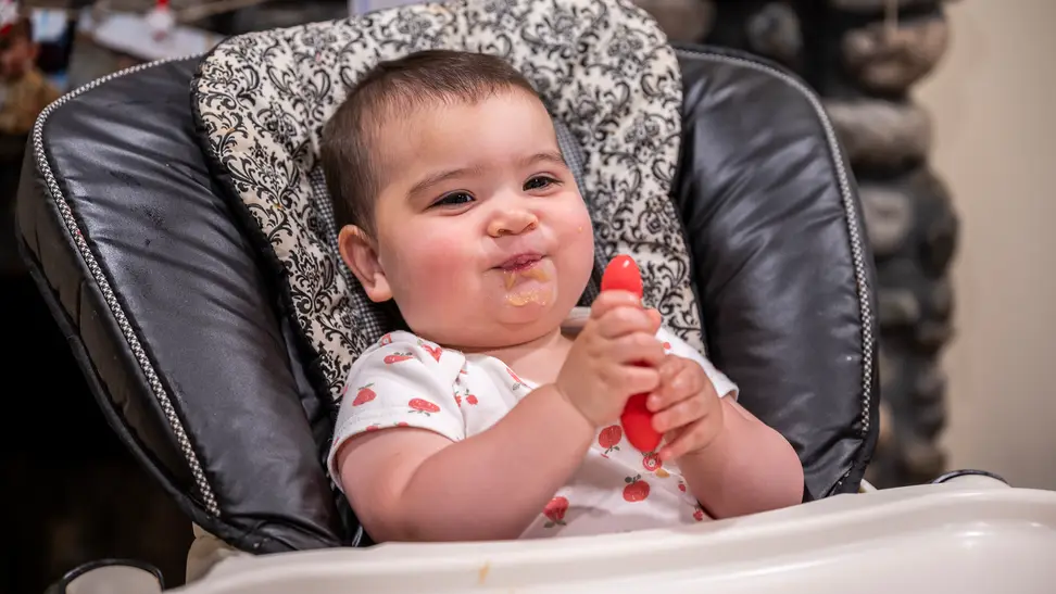 A baby in a high chair holding a baby spoon and smiling