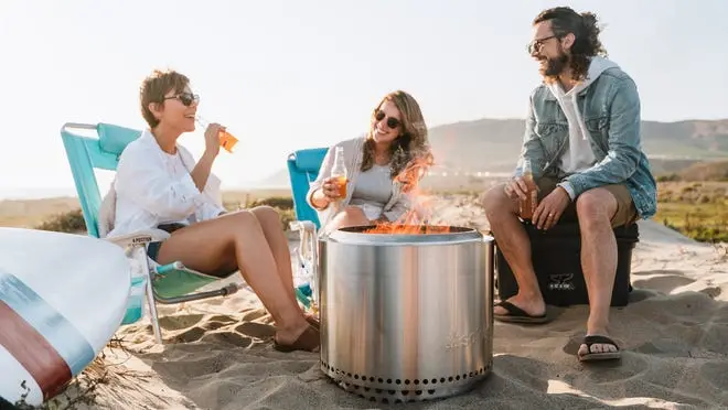 three people gathered around a solo stove fire on the beach