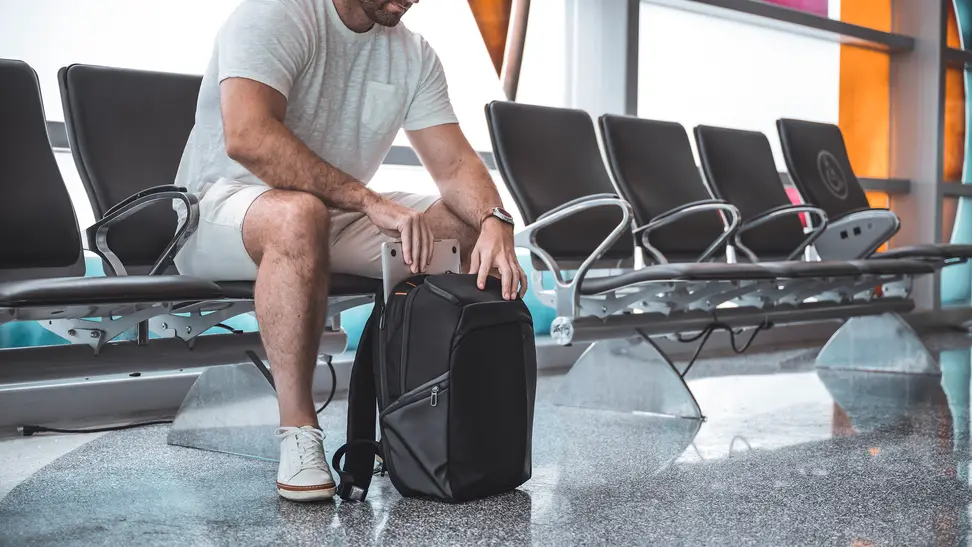 A man sits in an airport terminal, pulling a laptop out of a small backpack.
