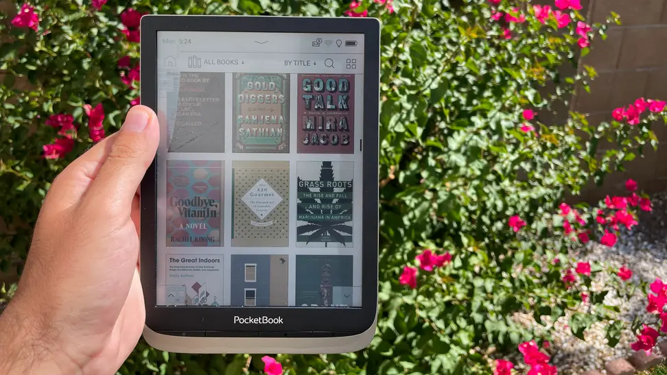 A hand holding an e-reader with a color screen against a backdrop with a large plant and pink flowers