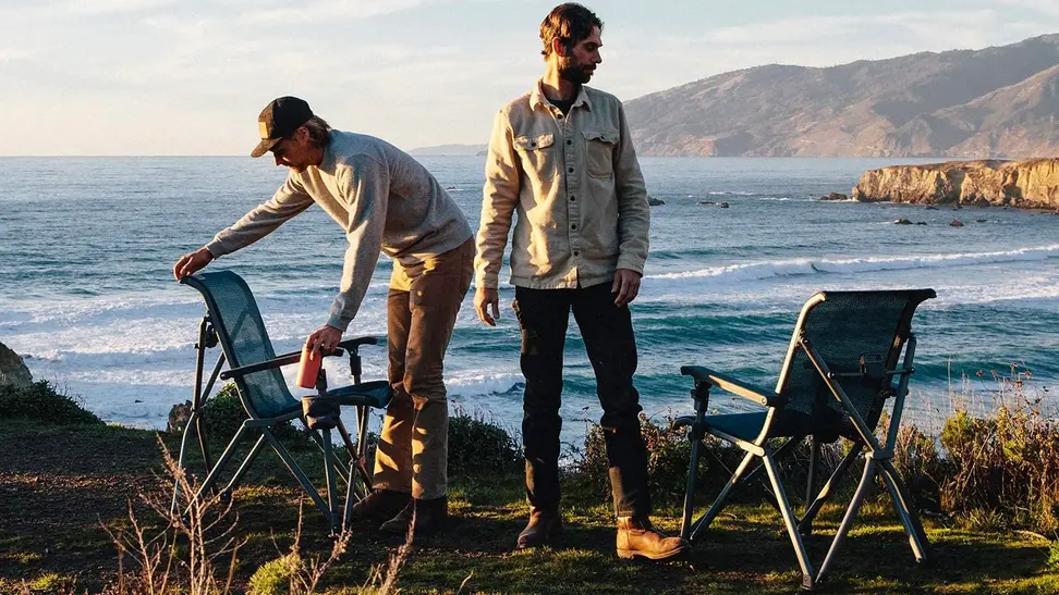 Two travelers setting their beach chairs up beside a large body of water.