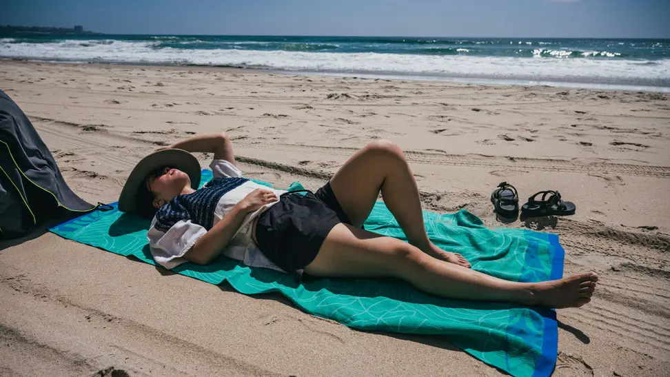 A woman reads a book while laying on a beach towel on the sand.