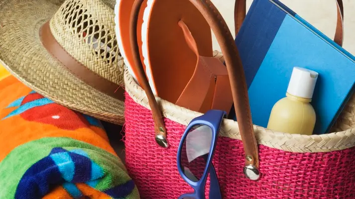 brightly colored beach accessories- towel, hat, flip flops, lotion, book, and sunglasses with a bag