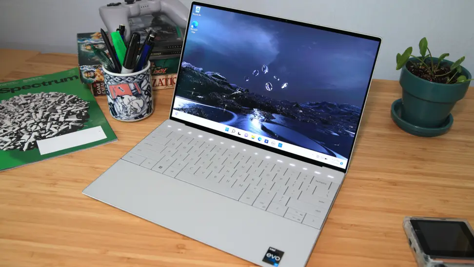 A silver laptop sits on a desk surrounded by a cup of pencils, a magazine, and a potted plant.
