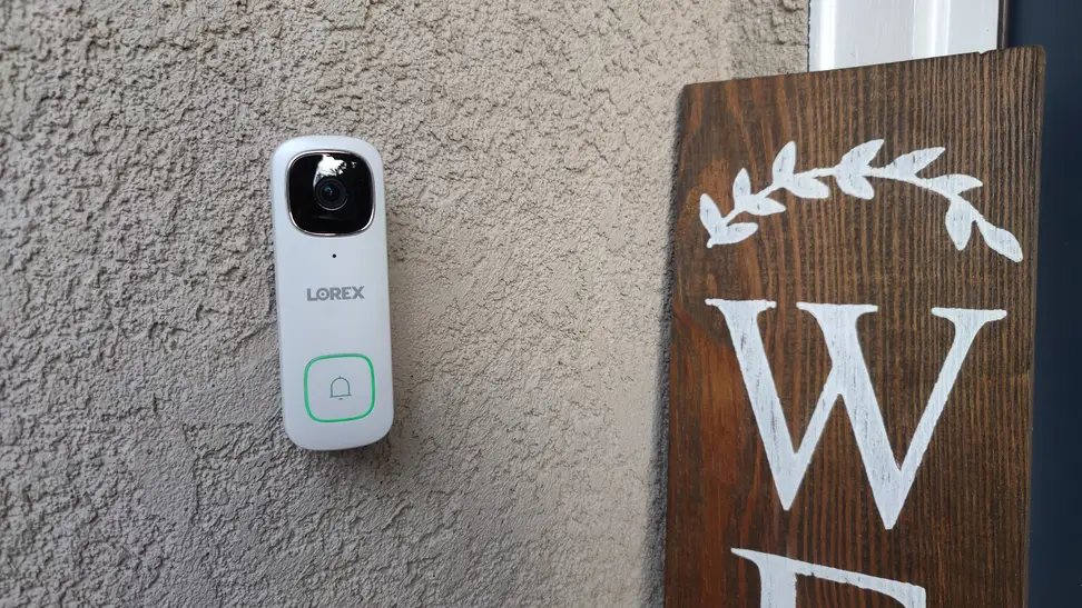 White Lorex 2K Wired Doorbell mounted on wall outside of home next to wooden welcome sign.
