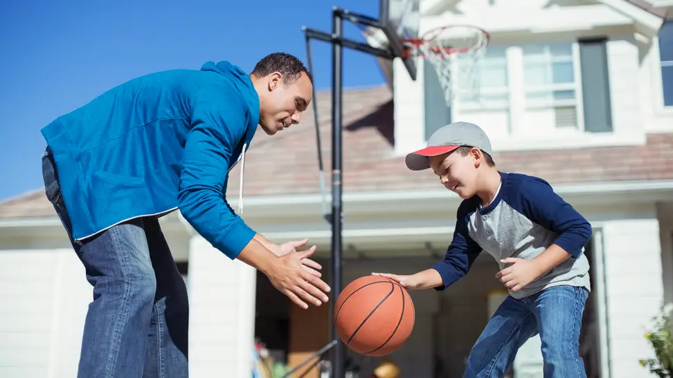 An adult and child play basketball in front of a house.