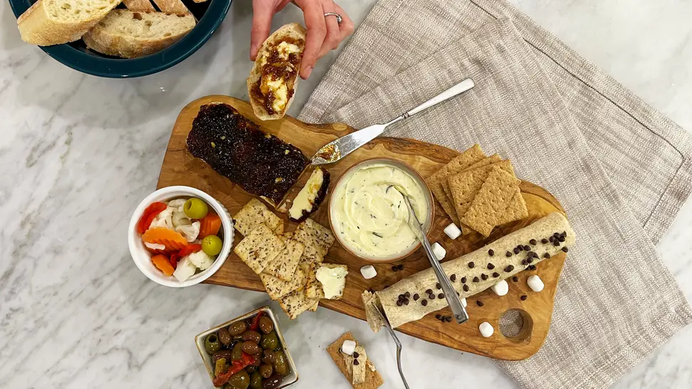 A hand holding a piece of bread with butter beside a butter board with accompaniments