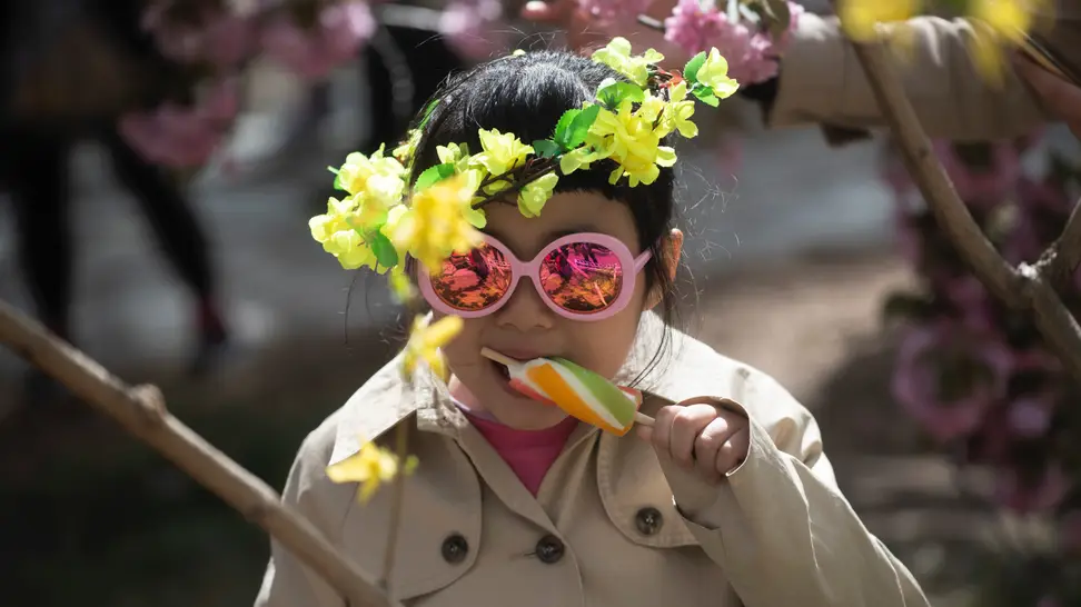 A child in pink sunglasses and a headpiece with yellow flowers enjoys a popsicle in the sun.