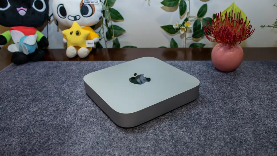 Apple Mac Mini M2 on top of a gray table mat with plushies and a small plant in the background