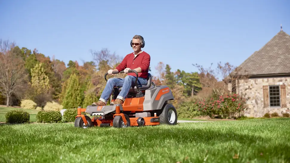 A person in a red shirt and jeans cuts their lawn using a Husqvarna zero-turn riding mower.