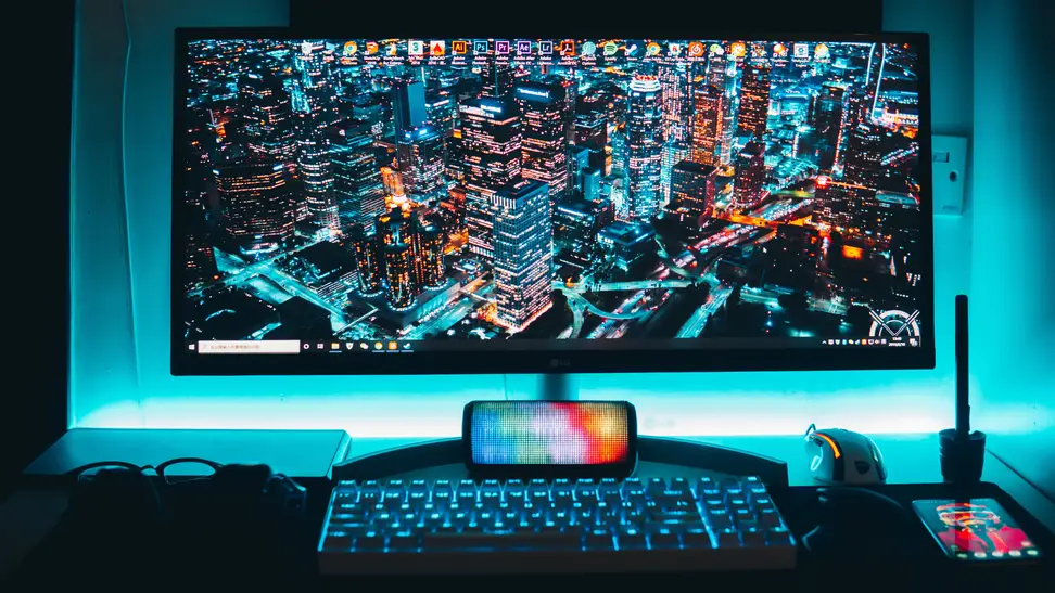 Desktop monitor with lit up back lights on top of desk surface behind keyboard with nighttime cityscape on screen.