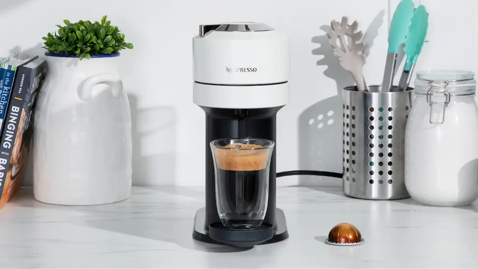Nespresso Vertuo coffee machine on marble countertop in the center of books, potted plants, utensils and sugar.