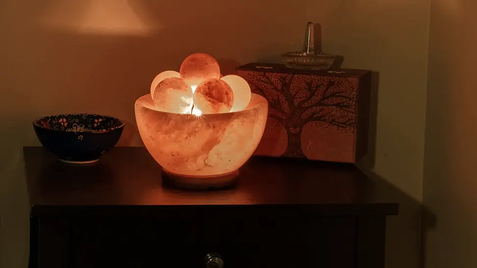 A glowing orange Spantik Himalayan Fire Bowl Salt Lamp glows on a wooden table. It has a number of crystalline massage balls inside.