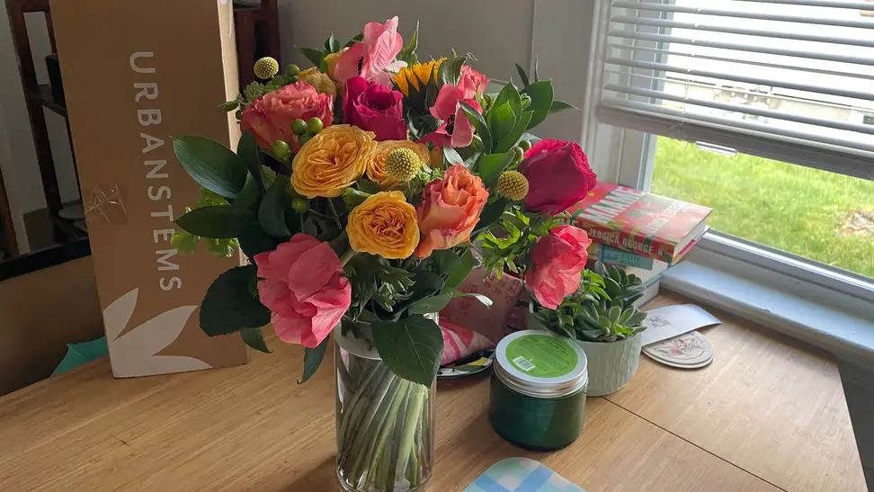 A bright bouquet of flowers from UrbanStems sitting on a wood table by a window
