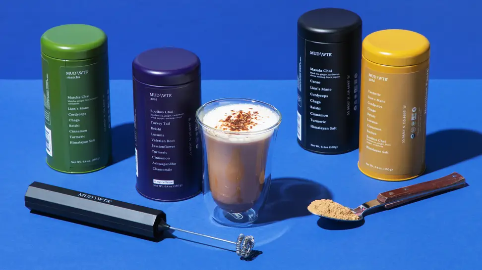 Mud/Wtr product containers next to a clear glass of a Mud/Wtr drink and a hand-held blender and spoon with Mud/Wtr product on it.