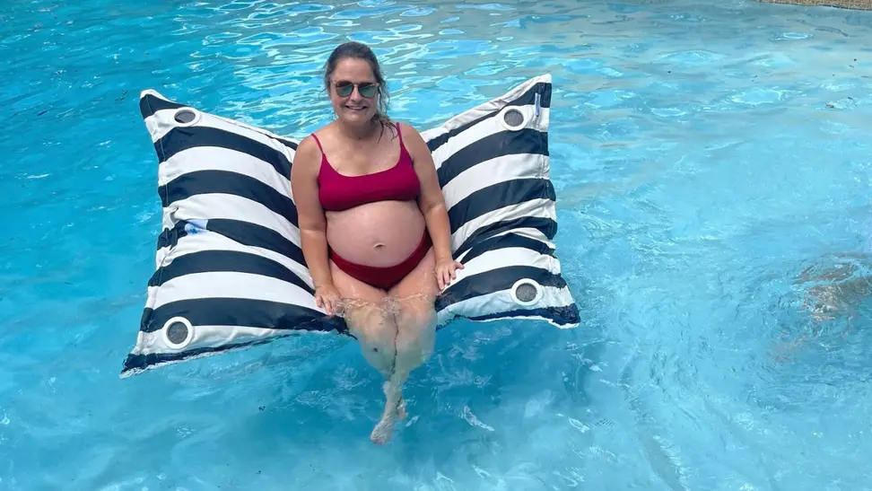 The Laze Pillow pool float in a fiddle leaf print floating in blue pool water with a woman sitting upright on it.