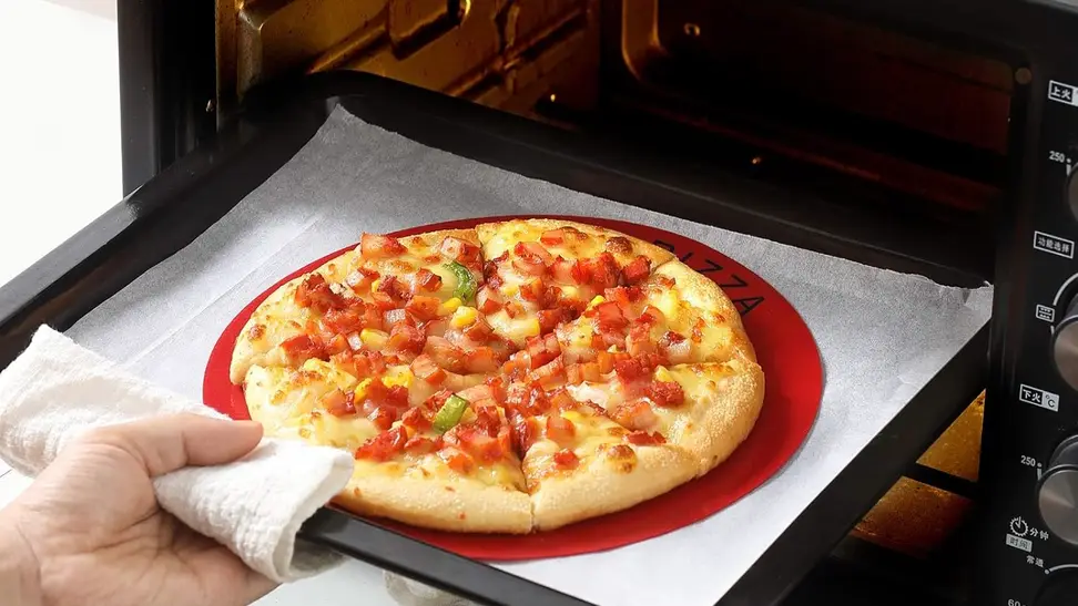 A pizza on top of a silicone baking mat is being placed in the oven.