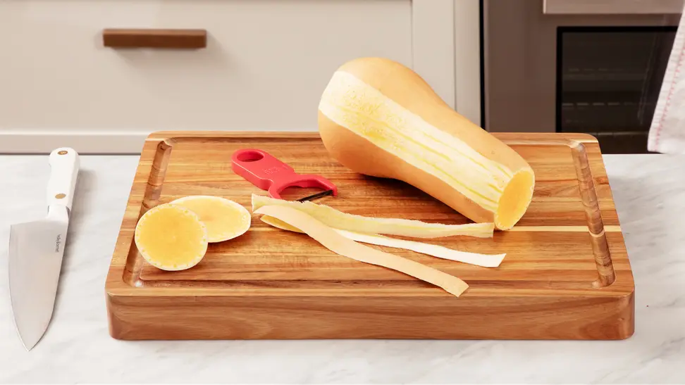 Peeled butternut squash with top and bottom parts removed sits on top of a wooden cutting board next to Y-peeler tool, knife and pile of peeled skin.