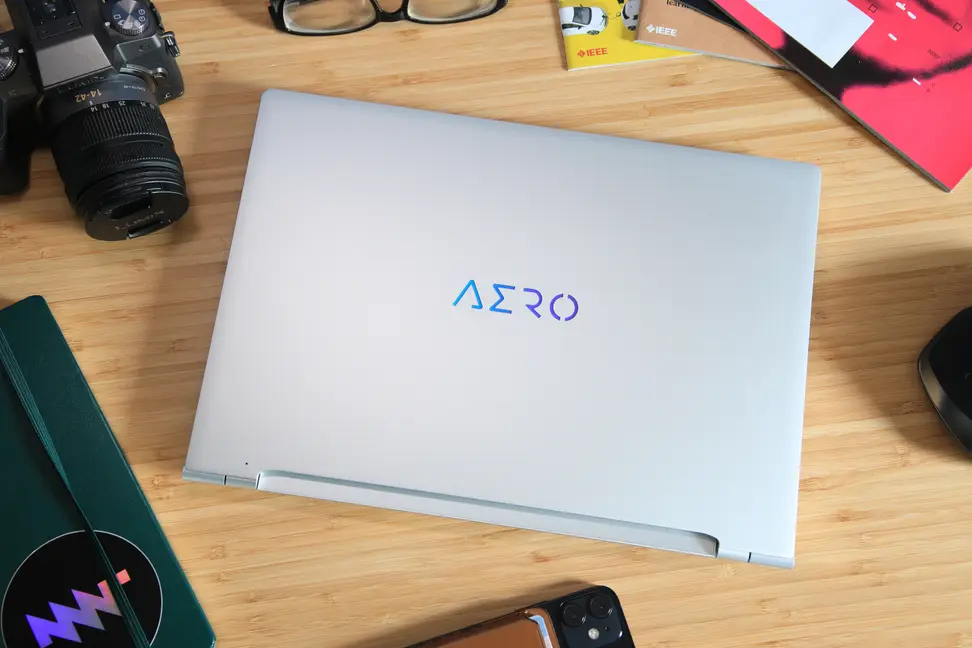 The Gigabyte Aero 14 OLED in silver, on a desk next to a camera and office supplies.