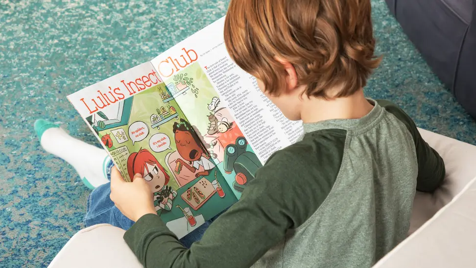 Child sitting in a small chair reading Highlights magazine.