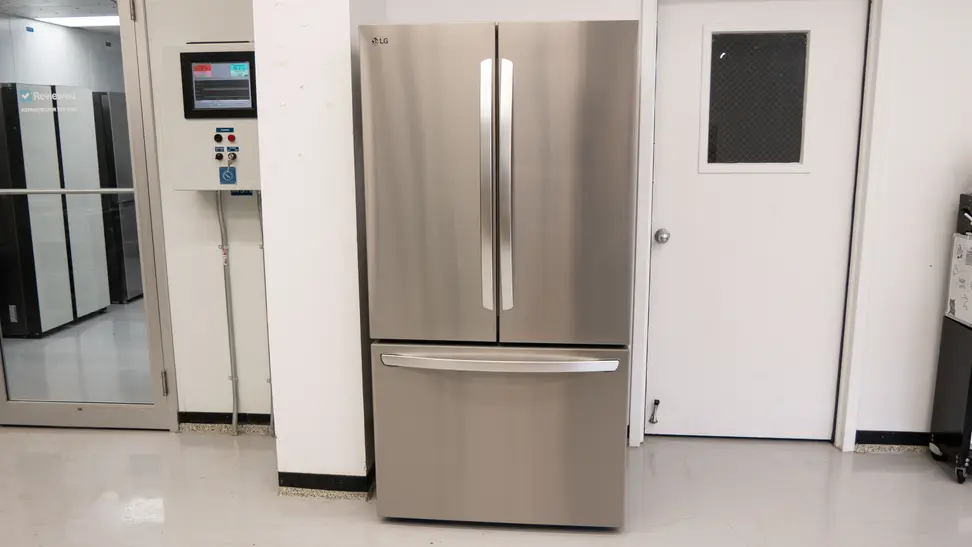 Front view of the silver LG LRFLC2706S Refrigerator against a wall in a white-walled product testing laboratory.