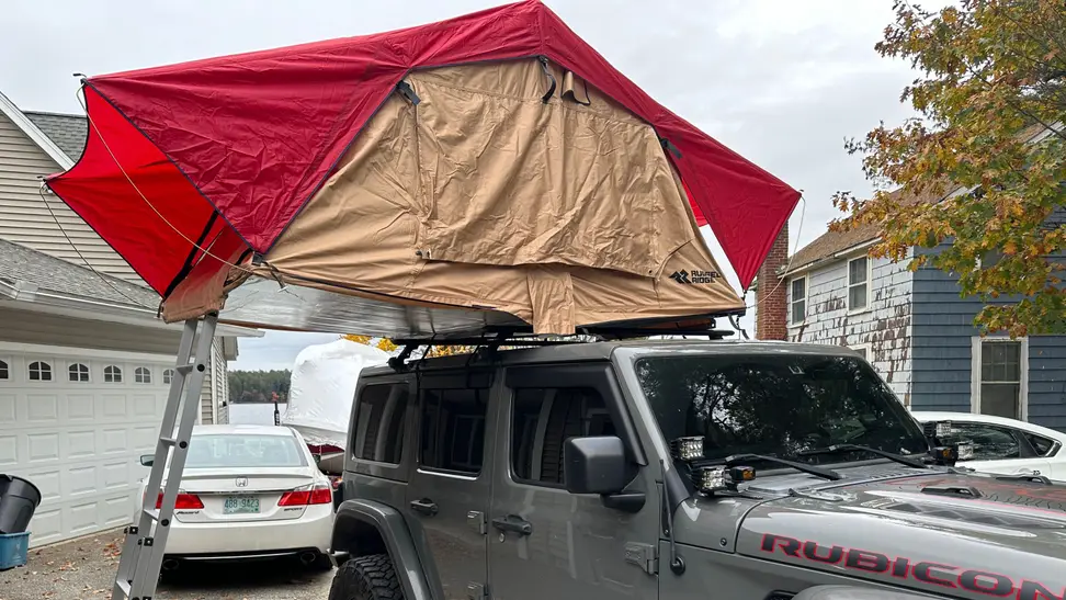 Angled front view of the Rugged Ridge Roof Top Tent set up over a gray Jeep Wrangler Rubicon in the front driveway of a house.