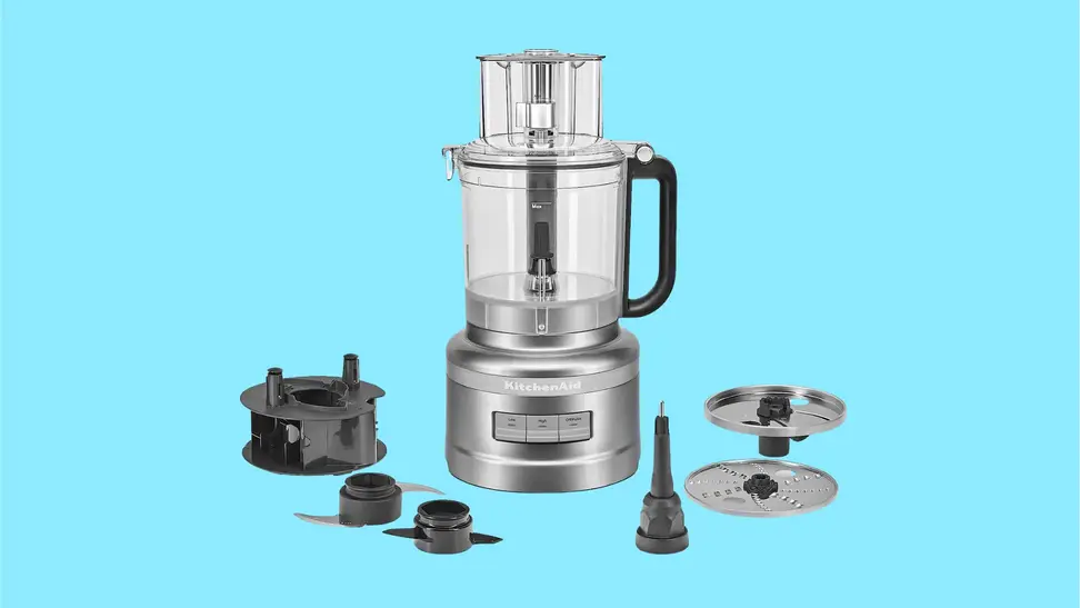 A KitchenAid 13-Cup Food Processor with its accessories on display in front of a colored background.
