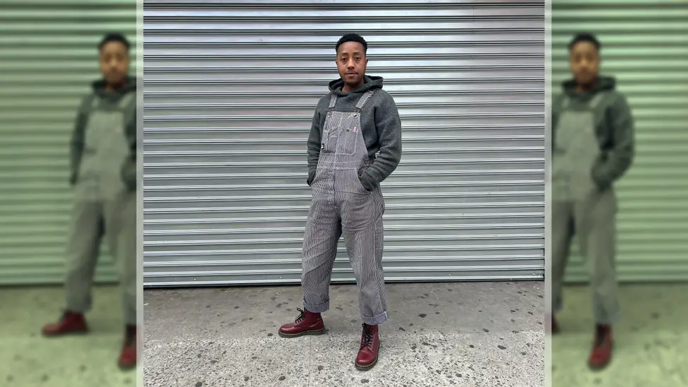 A person wearing overalls, red boots, and a gray hoodie, standing in front of a metal background.