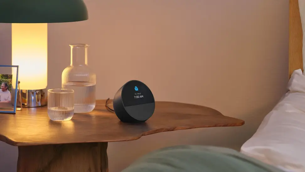 The Amazon Echo Spot smart alarm clock sits in a bedroom on a wooden nightstand.