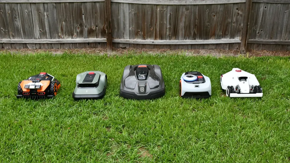 Five robot lawn mowers lined up together in the green grass with a fence behind