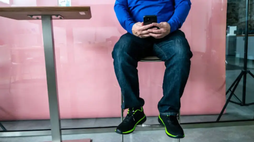 A person in jeans sits on a stool and is looking at their phone in front of a pink wall