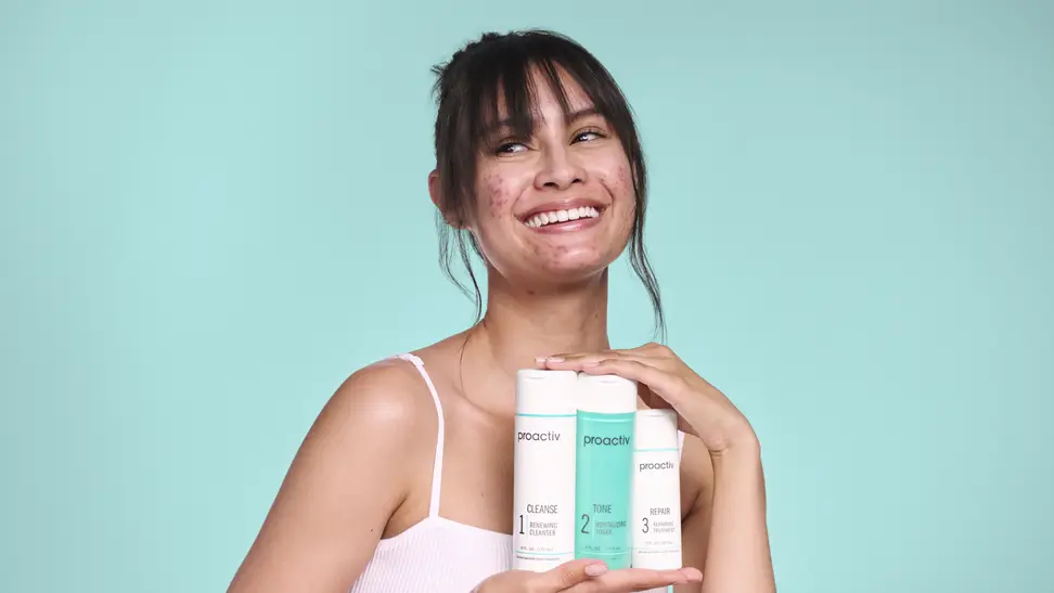A smiling young woman with acne holds up three proactiv acne treatment products against a teal background.