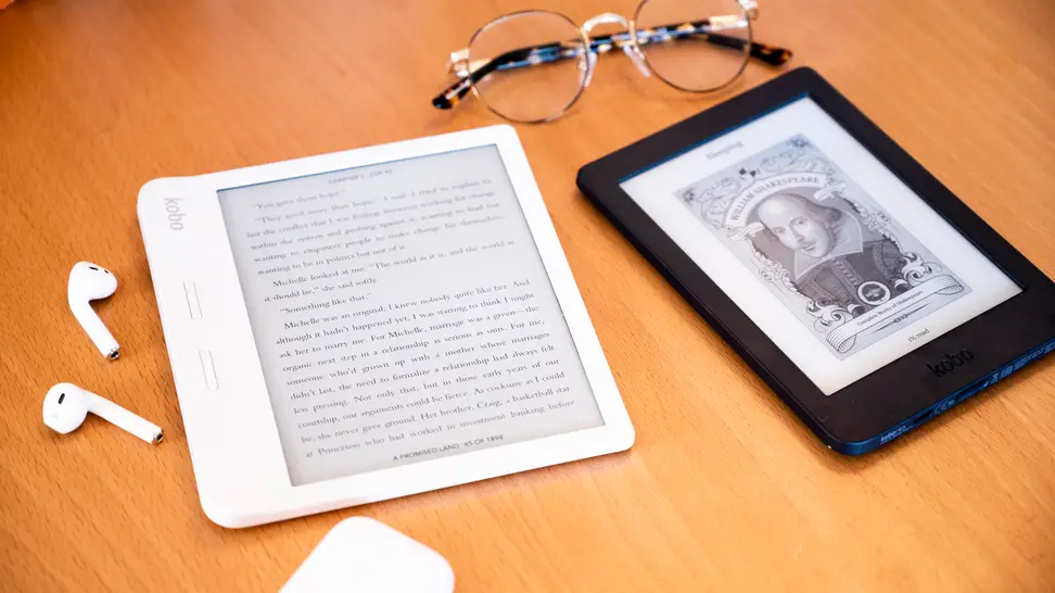 Two e-readers sitting on a wooden surface with a pair of glasses