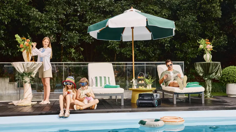 A young family relaxes by the pool under an umbrella while the Aiper Scuba X1 Pro Max cleans the pool nearby