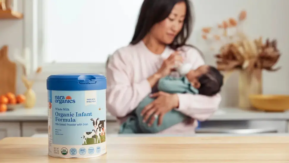 Mother holds baby and feeds with a bottle in the background while Nara Organics formula container sits on counter in foreground