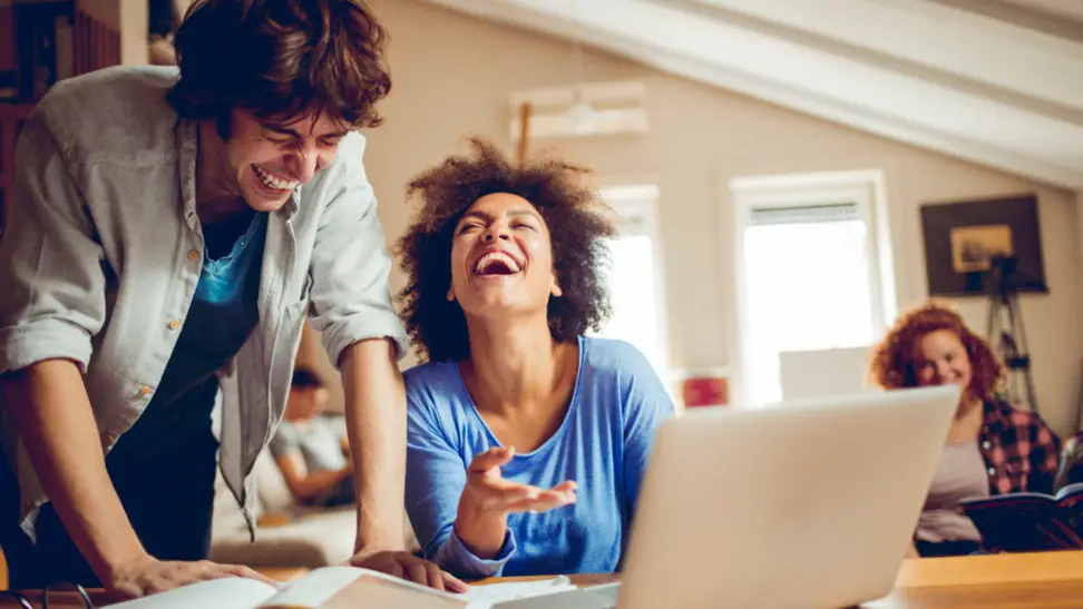 Two friends laughing together while working on a laptop at home, highlighting the fun and connectivity possible with T Mobile 5G Home Internet.