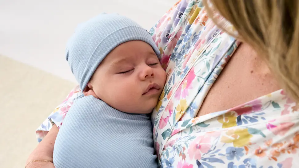 A newborn baby swaddled in blue, sleeping peacefully in a caregiver’s arms, promoting comfort and savings with a little sleepies coupon code.