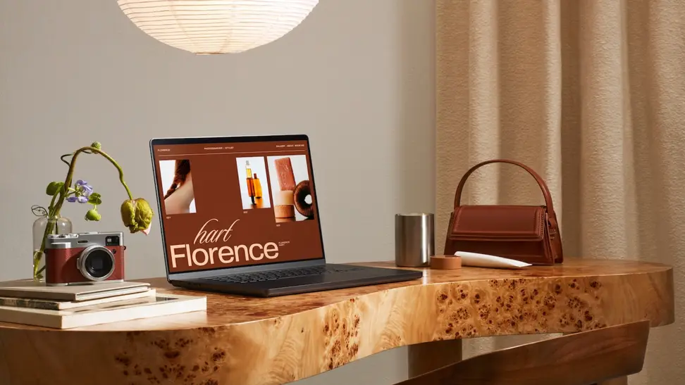 A stylish workspace with a laptop showcasing a modern Squarespace templates design on a wooden desk beside a camera, vase, and leather bag.