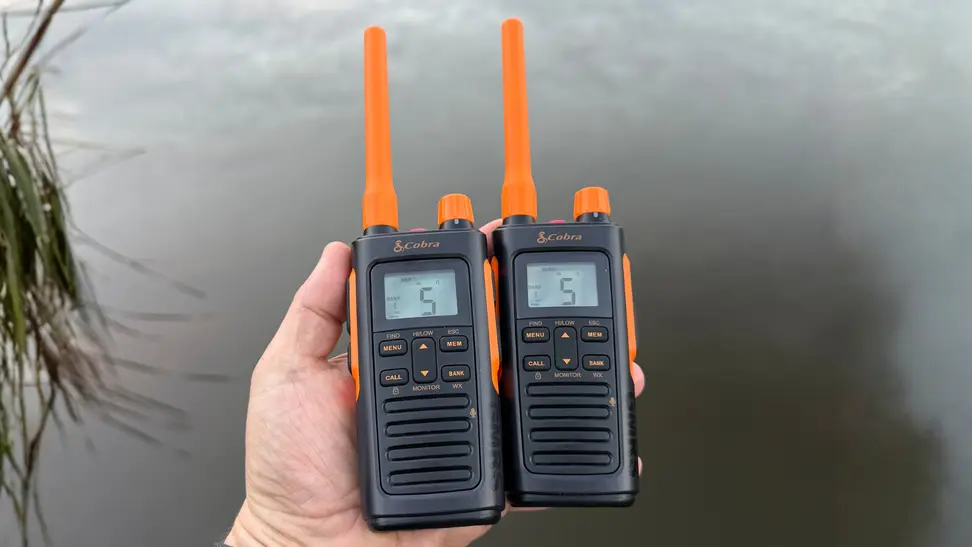 Two Cobra walkie-talkies held in hand with a lake in the background