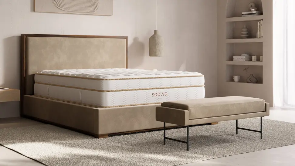 A modern bedroom featuring a white Saatva Classic mattress on a low-profile upholstered bed frame with warm wood accents, styled with neutral decor and soft natural lighting—an image highlighting the Saatva Memorial Day sale on luxury mattresses.
