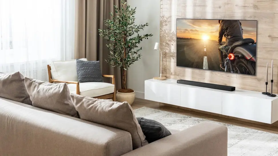 A bright, modern living room featuring an LG Dolby Atmos soundbar mounted beneath a large flat-screen TV on a light wood accent wall. The minimalist space includes a beige sectional sofa, white media console, indoor plant, and soft neutral decor, showcasing a clean and immersive home entertainment setup.