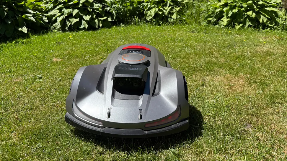 Silver Sunseeker Elite X7 robot lawn mower on grass in front of some bushes.